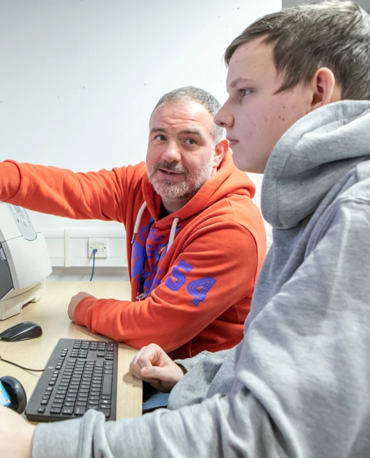 Lecturer in an orange hoodie guiding a student through programming a robotic vehicle on a computer screen in a computing classroom. Lecturer in an orange hoodie guiding a student through programming a robotic vehicle on a computer screen in a computing classroom.